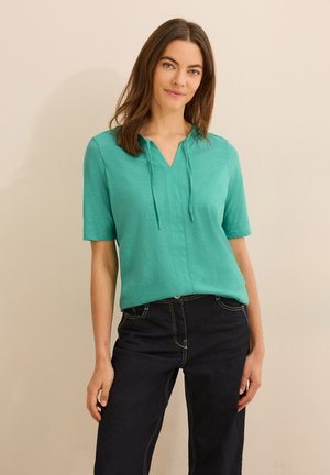 Haut turquoise à manches courtes avec décolleté en V et détail à cordon, associé à un jean en denim foncé avec des coutures visibles et des poches.