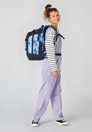 Teenagermädchen trägt lavendelfarbene Overalls und gestreiftes Shirt, trägt großen blauen Rucksack mit Muster, lächelt, steht vor einfachem Hintergrund.
