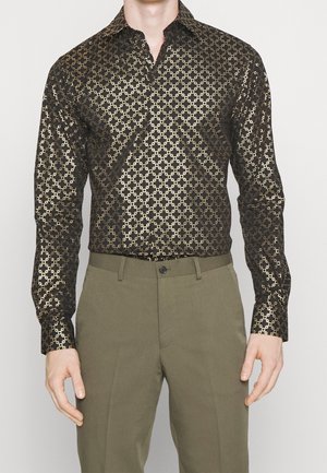 Mann trägt dunkles Langarmhemd mit goldenem geometrischem Muster und olivgrüner Hose, steht vor einfarbigem Hintergrund.