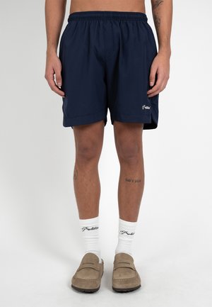 Uomo che indossa pantaloncini blu navy, calzini bianchi con marchio e scarpe beige senza lacci, in piedi davanti a uno sfondo bianco semplice.