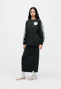 Μαύρο φούτερ με λευκές ρίγες, λουλουδάτο έμβλημα στο στήθος· συνδυασμένο με μαύρη μακριά φούστα με φερμουάρ στο πλάι. Το μοντέλο φοράει ελαφριά υποδήματα.