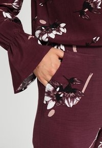 Conjunto color burdeos con patrones florales en blanco y negro; presenta mangas acampanadas y un bolsillo. La tela parece suave y texturizada.