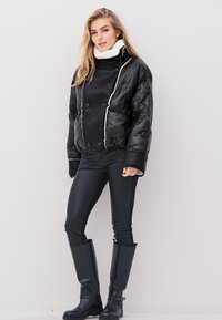 Fekete pufferszövet kabát fehér részletekkel, vastag gallérral és fényes textúrával. Fekete, testhezálló nadrággal és térd fölé érő fekete csizmával párosítva.