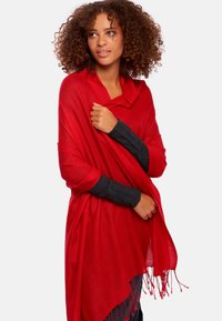 Écharpe rouge avec des bords frangés, fabriquée en tissu doux. Elle présente un devant ouvert et des manches larges, superposée sur un haut ajusté sombre.