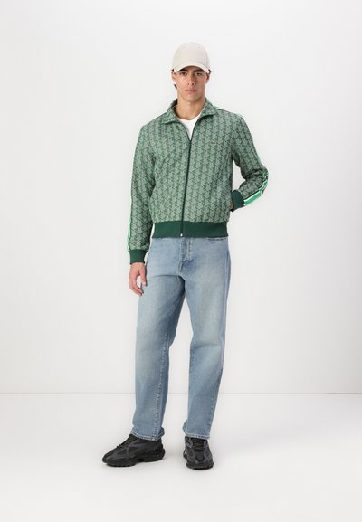 Veste bomber zippée verte en tissu à motifs, manches rayées contrastantes, poignets élastiques, associée à un jean bleu clair et des baskets noires.