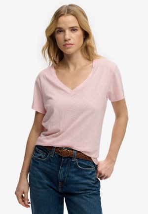Donna bionda che indossa una t-shirt rosa chiaro con scollo a V infilata nei jeans blu con cintura intrecciata marrone, in piedi con una mano in tasca.
