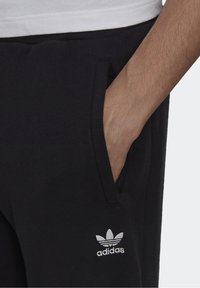 Svarta sweatpants i mjukt tyg, med sidofickor och en liten vit Adidas-logotyp på vänster lår. Elastisk midja ingår.