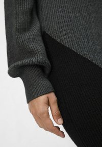 Maglione a maglia grigio scuro con una texture a coste, caratterizzato da un motivo diagonale e una sezione nera a contrasto. Mano visibile in posizione rilassata.