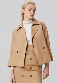 Veste courte beige avec un large col, des doubles boutons et des manches trois-quarts; assortie à une jupe coordonnée avec une ceinture.