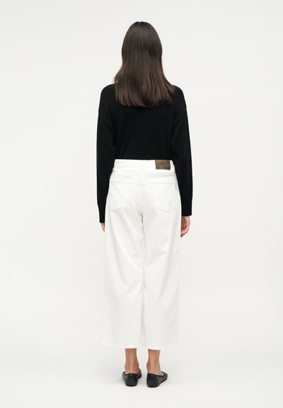 Άτομο με μακριά καστανά μαλλιά που φοράει μαύρο μακρυμάνικο τοπ, λευκό παντελόνι με φαρδιές ρεβέρ και μαύρα παπούτσια slip-on, στέκεται με την πλάτη προς τα εμπρός.