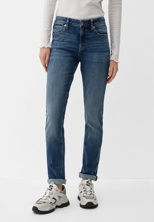 Person in hochgekrempelten blauen Jeans, weißem, geripptem Langarmshirt und weißen klobigen Sneakers steht vor einem schlichten Hintergrund.