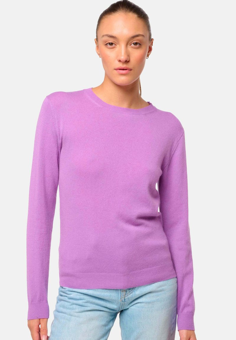 Femme portant un pull long à manches longues violet clair et un jean bleu clair, se tenant devant un fond uni clair.
