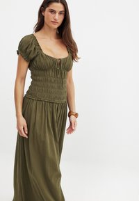 Olivgrünes, gesmoktes Maxikleid mit kurzen puffärmel, quadratischem Ausschnitt und Schlüsselbeindetail auf der Vorderseite. Strukturiert Stoff.
