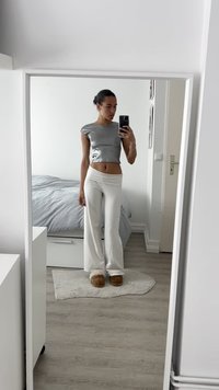 Srebrny top z krótkim rękawem połączony z jasnobeżowymi spodniami o szerokich nogawkach i brązowymi klapkami. Odbijający materiał i gładka faktura tkaniny. Minimalistyczne otoczenie.