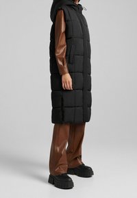 Gilet matelassé noir avec capuche, présentant une texture matelassée et des poches latérales, associé à un pantalon en simili cuir marron et des bottes à plateforme noires chunky.