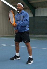 Jogador de ténis segurando uma raquete laranja, vestindo um capuz azul com o texto "Kale Court", calções pretos e sapatilhas Nike pretas, num campo.