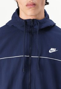 Mornarica zip-up jakna s kapuco, bela nagibna črta čez prsa in Nike logotip na levi strani. Narejena iz lahkega materiala.