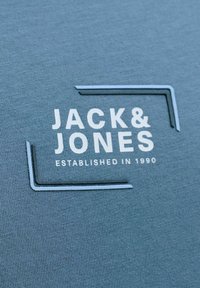 Tela azul con estampado gráfico blanco que muestra el texto "JACK & JONES" y "ESTABLISHED IN 1990", enmarcado por un contorno rectangular.