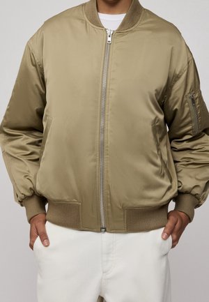 Personne portant une veste bomber marron clair fermée par un zip, avec des poignets et un ourlet côtelés, les mains posées négligemment dans les poches d’un pantalon blanc.