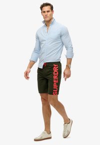 Groene zwembroek met rood "SUPERDRY" logo, met een trekkoord in de taille. Lichtblauw shirt met lange mouwen en knoopdetails. Beige sneakers.