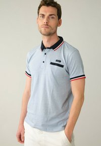 Světle modré polo tričko s námořnickým límcem a červenými akcenty, vybavené kapsou na hrudi a dvěma knoflíky na krku. Krátké rukávy. Bavlněný materiál.