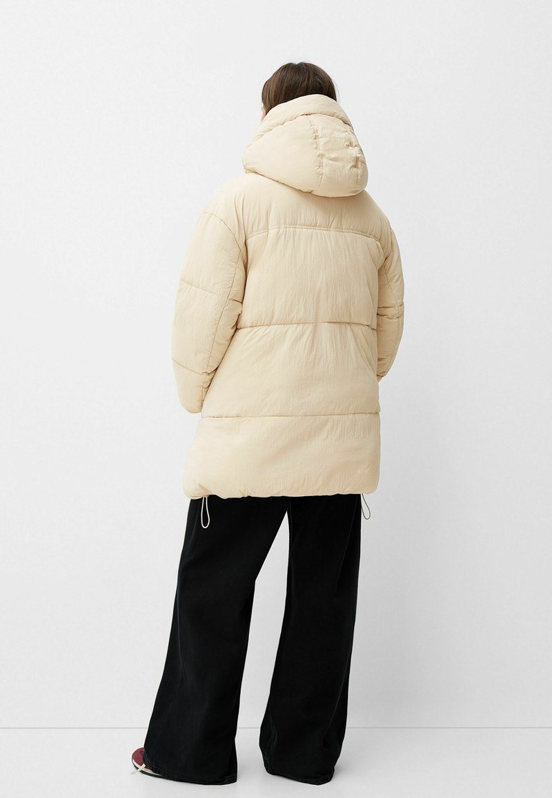 Giubbotto imbottito beige con cappuccio, caratterizzato da un design trapuntato e coulisse elastiche all'orlo. Abbinato a pantaloni neri a gamba larga.