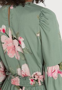 Robe florale verte avec des fleurs roses, dotée d'une ouverture en forme de goutte dans le dos, de manches bouffantes et d'une taille smockée. Confectionnée dans un tissu doux et léger.