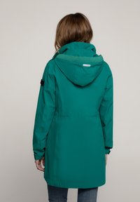 Türkisfarbene wasserdichte Jacke mit Kapuze, elastischen Bündchen und verstellbarem Bund. Sie hat ein strukturiertes grün gefüttertes Innenfutter und einen reflektierenden Akzent.