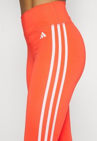 Höga leggings i lysande orange tyg med tre vita ränder längs sidan och en liten vit logotyp på midjebandet.