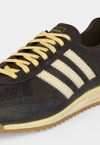 Σκούρο καφέ αθλητικό παπούτσι Adidas με υφή στην επιφάνεια, κρεμ τρεις ρίγες, κρεμ κορδόνια και λαστιχένια σόλα με λογότυπο Adidas στο πλάι.
