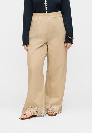 Kvinde iført beige vidde bukser med bølgede, hulmønstrede blonder i kanten, kombineret med en navy bluse med lange ærmer og brune sandaler.