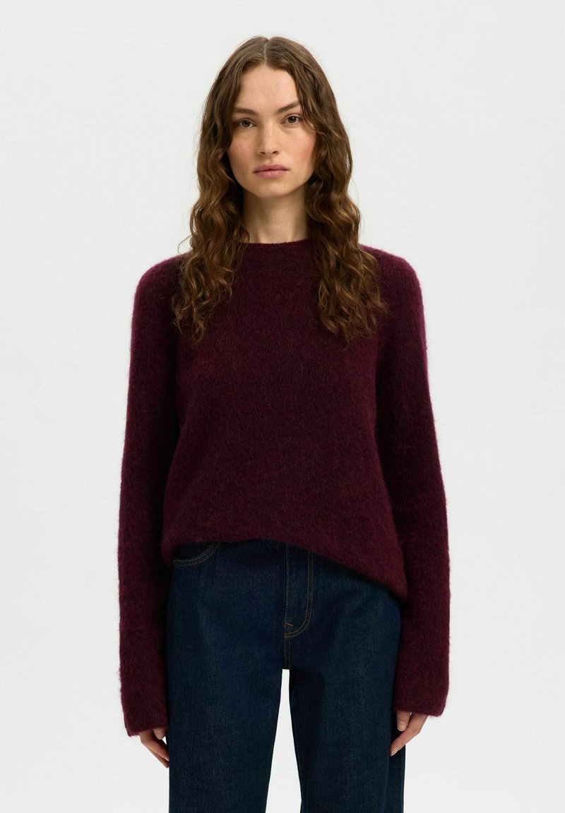 Donna con capelli lunghi e mossi, che indossa un maglione morbido viola scuro e jeans blu scuro, in piedi davanti a uno sfondo chiaro e semplice.