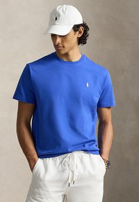 Man wearing blue short-sleeve shirt, white cap, and white drawstring pants with hands in pockets against neutral background.