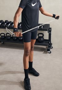 Camiseta atlética negra con un logo blanco, pantalones cortos de compresión negros, calcetines negros y zapatos; sosteniendo una banda de resistencia; pesas de fondo.
