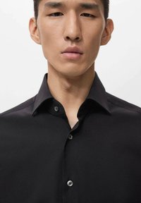 Camisa negra de botones con collar estructurado y textura suave, que presenta cuatro botones grises en la tapeta y un diseño ajustado.