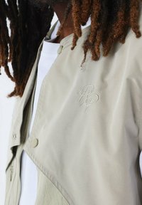 Giacca beige chiaro con una texture liscia, dotata di una chiusura parziale a scatto e logo ricamato sul petto. Camicia bianca sottostante visibile.