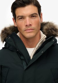 Ciemnozielona parka z kapturem wyłożonym futrem, z zamkiem błyskawicznym, napami oraz miękką kremową podszewką.