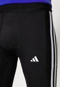 Schwarze Leggings aus dehnbarem Stoff, mit einem weißen Dreistripedesign an der Seite und einem weißen Logo am unteren Bein.