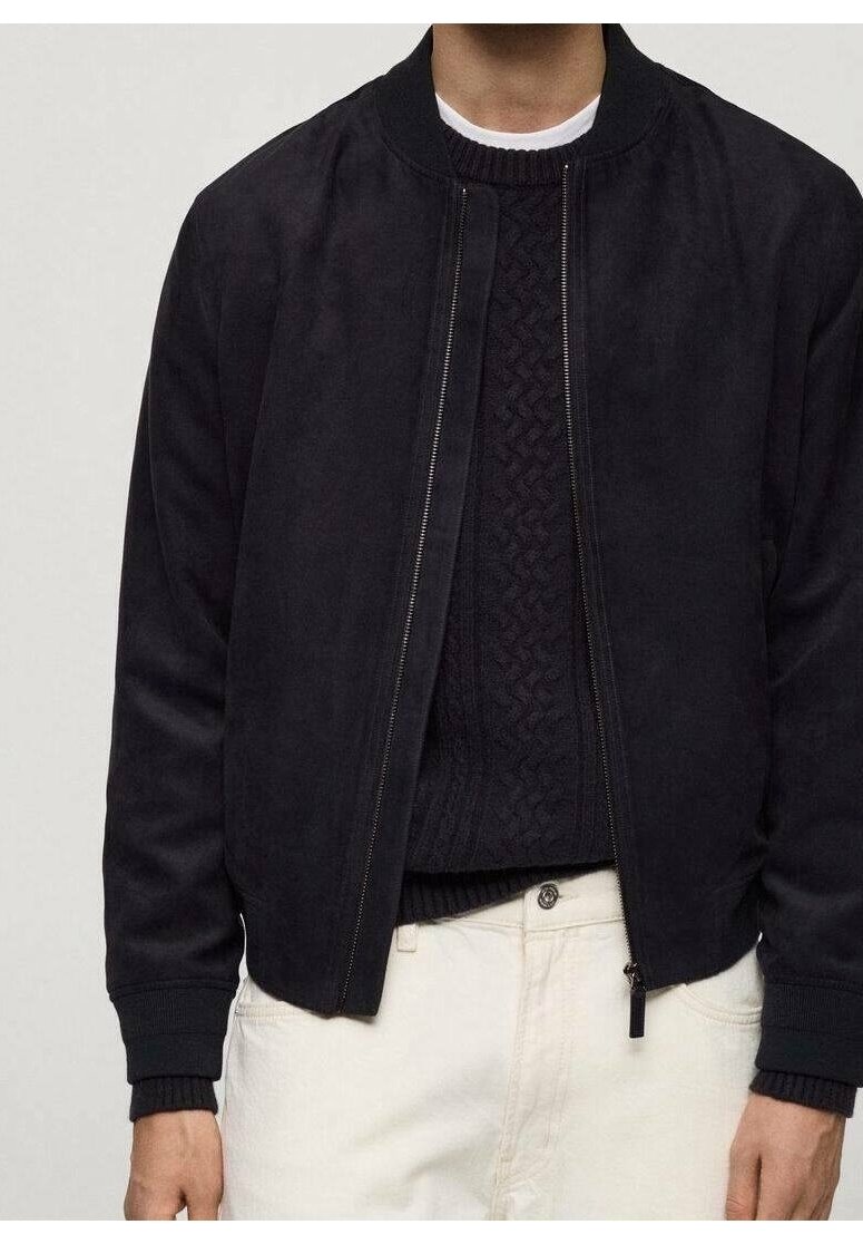 Personne portant un blouson bomber noir zippé sur un pull en maille torsadée noir et un pantalon blanc, debout devant un fond uni.