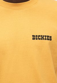 Κίτρινη βαμβακερή μπλούζα με στρογγυλή λαιμόκοψη και την λέξη "DICKIES" τυπωμένη με έντονα μαύρα γράμματα στην αριστερή πλευρά.