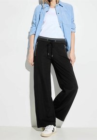 Svarta vida byxor med dragsko i midjan, med sido-fack med dragkedja och en mjuk tygkvalitet. Matchat med en ljusblå skjorta.