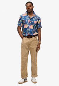 Korte mouwen blauwe shirt met kleurrijke bloemen- en abstracte patronen, gecombineerd met beige broek en witte sneakers met zwarte accenten.