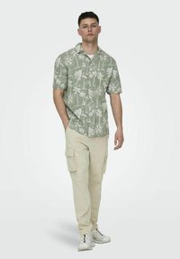 Korte mouwen groene shirt met een wit tropisch patroon; gecombineerd met beige cargobroek. Model draagt witte sneakers en staat neutraal.