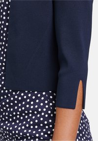 Marinblå stickad cardigan med trekvartslånga ärmar, med en sidoklyfta. Underneath en klänning med prickigt mönster i marinblått och vitt.