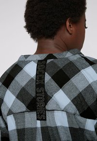 Camicia a quadri nera e blu chiaro con un tessuto strutturato. Presenta una toppa sul retro con il testo "ROCK STORIES," abbellita con strass.