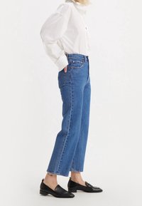 Vaqueros de mezclilla azul con un diseño de pierna recta. Cintura alta, estilo clásico de cinco bolsillos y detalles de costura en contraste. Llevado con una camisa blanca y mocasines negros.