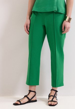 Persona que lleva pantalones recortados de color verde brillante, sandalias planas negras con tiras y una pulsera de brazalete dorada en la muñeca izquierda sobre un fondo liso.