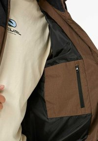 Bruine jas met een zwart interieur, voorzien van een ritszak. Begeleid door een beige T-shirt met een blauw logo.