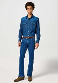Blaue Jeanshemden mit zwei Brusttaschen, Knopfkragen und dekorativer Stickerei. Figurnah geschnittene blaue Jeans und brauner Gürtel. Braune Cowboy-Stiefel.