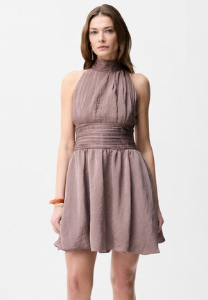 Kvinde iført ærmeløs mauve halterneck-kjole med plisseret taljebånd, stående mod en ensartet lys baggrund, iført armbånd på venstre håndled.